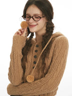 Close-up of a woman's face wearing a brown cable knit hooded sweater with buttons and pom-poms, playing with the pom-poms.