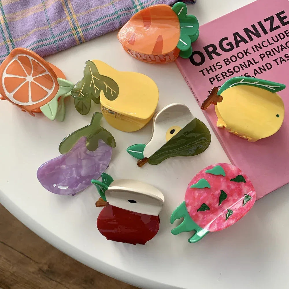 Top-down view of fruit-shaped hair claw clips scattered on a white surface with a pink book in the background