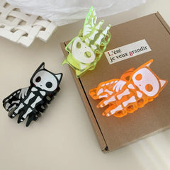 Three skeleton cat claw hair clips in black, orange, and neon green on a box