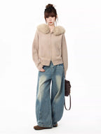 Full body shot of a woman wearing a beige knit cardigan with a faux fur collar and wide-leg blue jeans, holding a brown backpack