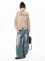 Full body shot of a woman wearing a beige knit cardigan with a faux fur collar and wide-leg blue jeans, holding a brown backpack