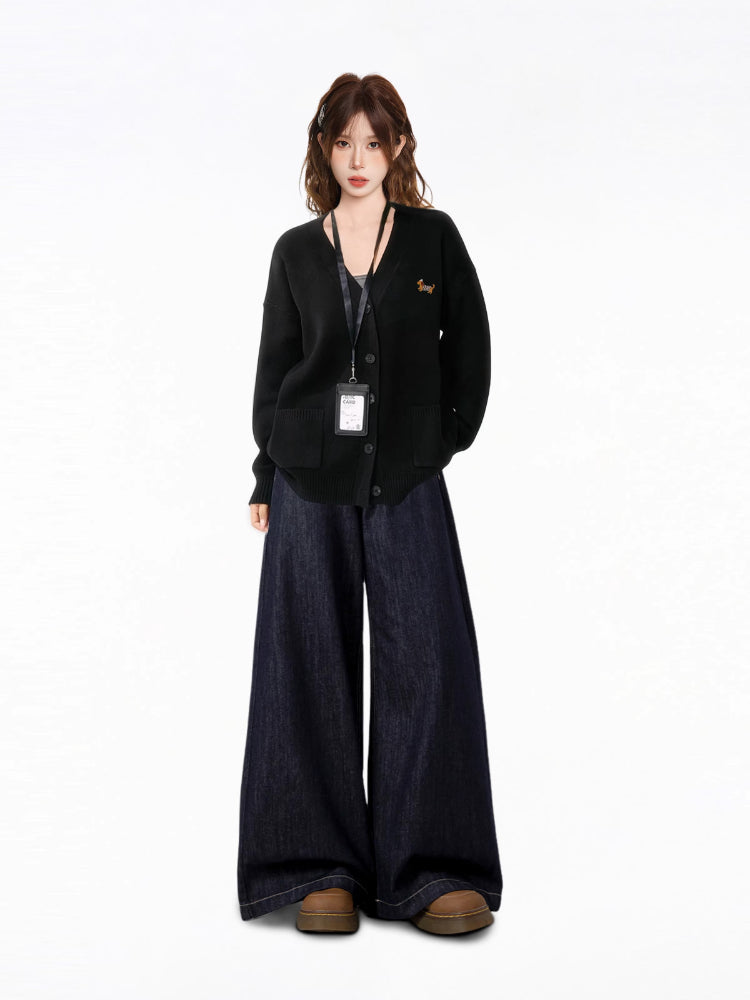 Full body shot of a model wearing a black V-neck knit cardigan with an embroidered dachshund detail, a black tank top, and dark wash wide-leg jeans, with an ID badge holder around her neck.