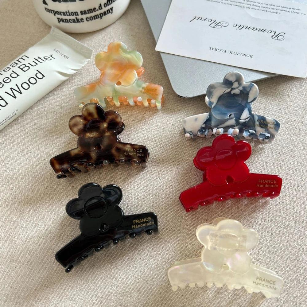 A variety of flower-shaped claw clips are displayed on a neutral background, showcasing different colors and marbled patterns. Text is visible on some clips: "FRANCE Handmade".