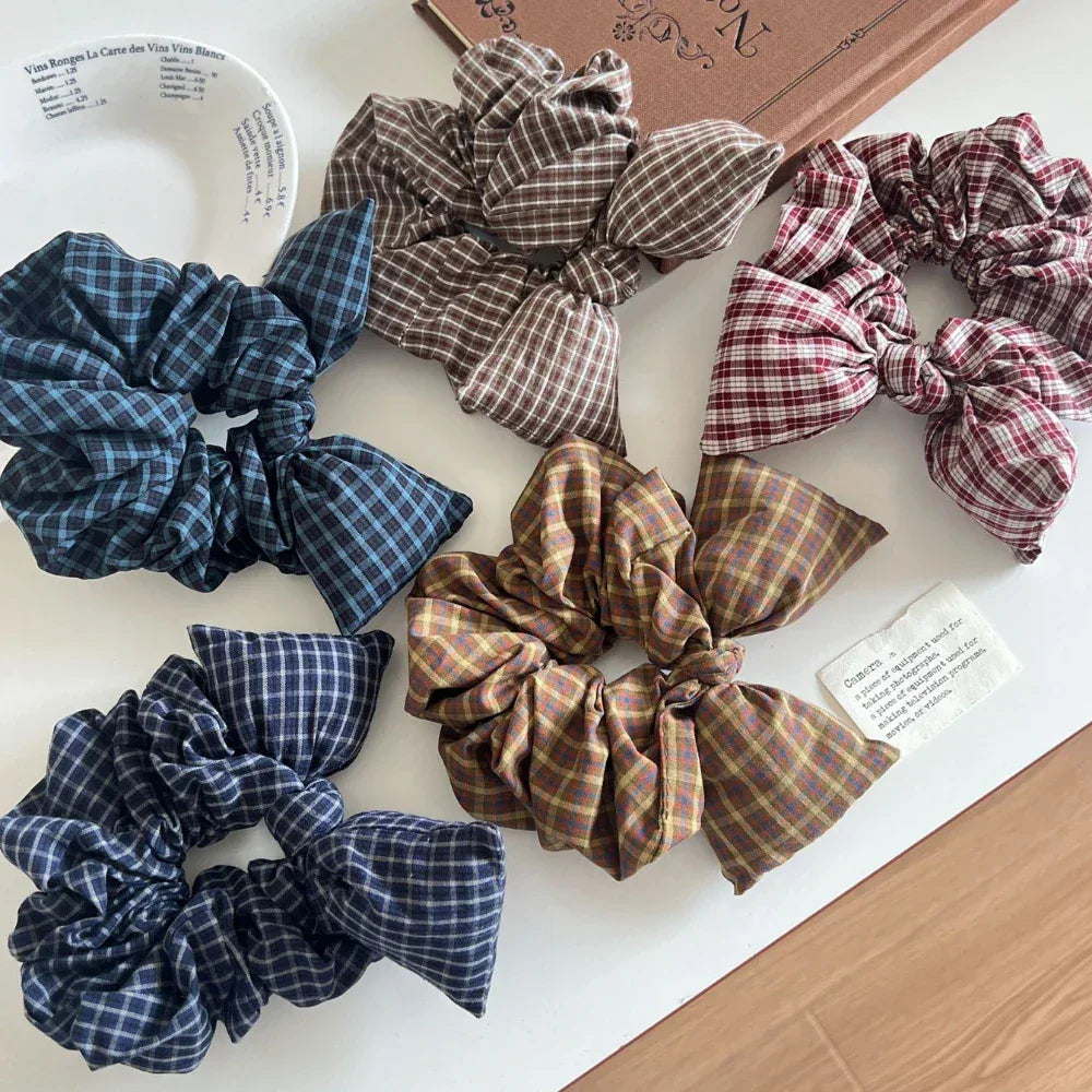 Several plaid scrunchies with bow details are displayed on a white tabletop, some partially in a tray.