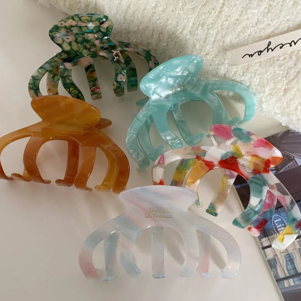 A group of six acetate claw hair clips in various marbled colors: green, light blue, amber, multi-colored, and white with pink/blue accents. They are arranged on a white textured surface.