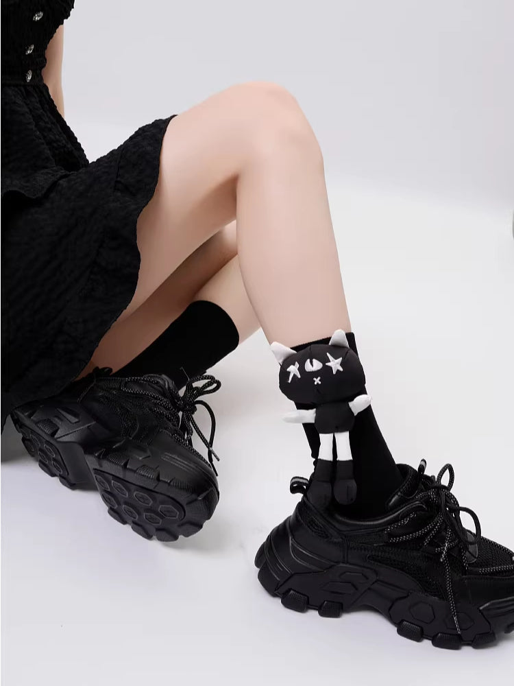 A person is sitting down wearing black socks with cat appliques and black platform shoes.