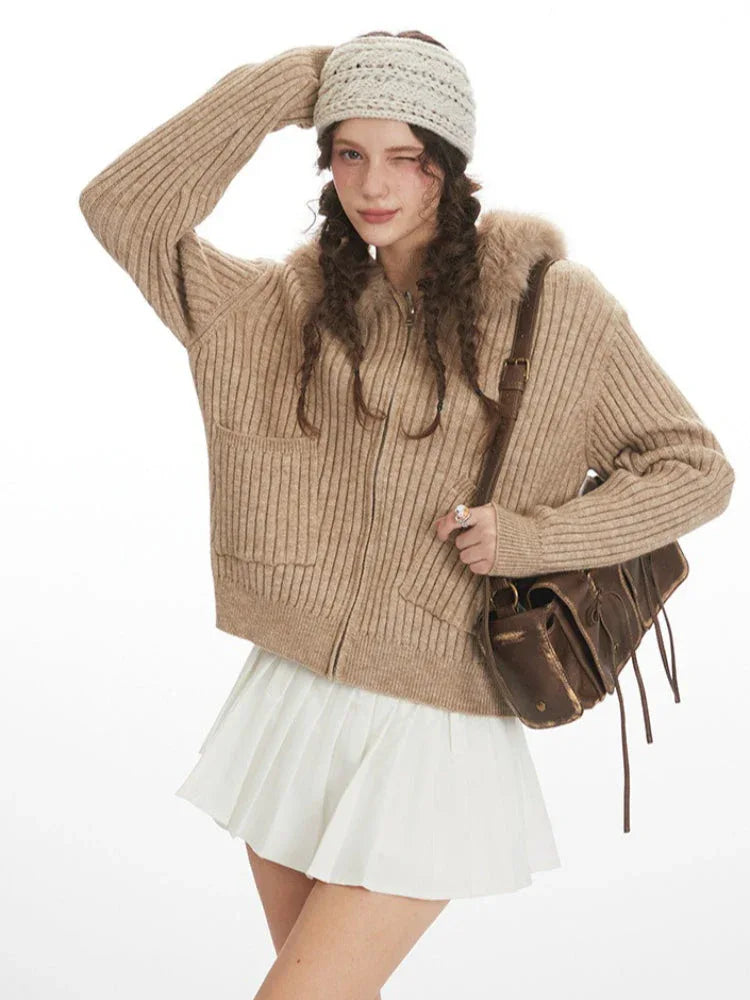 Close-up of a model wearing a beige ribbed knit hooded jacket with faux fur trim, winking and touching her hair.