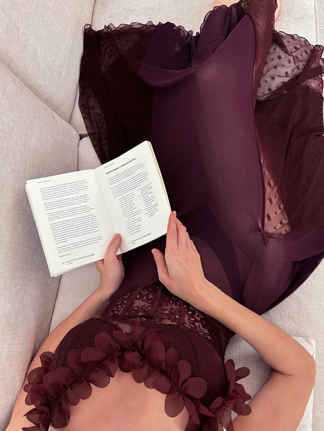 Woman wearing a sheer plum maxi slip dress with floral applique straps and lace bodice, lying on a cream sofa reading a book.