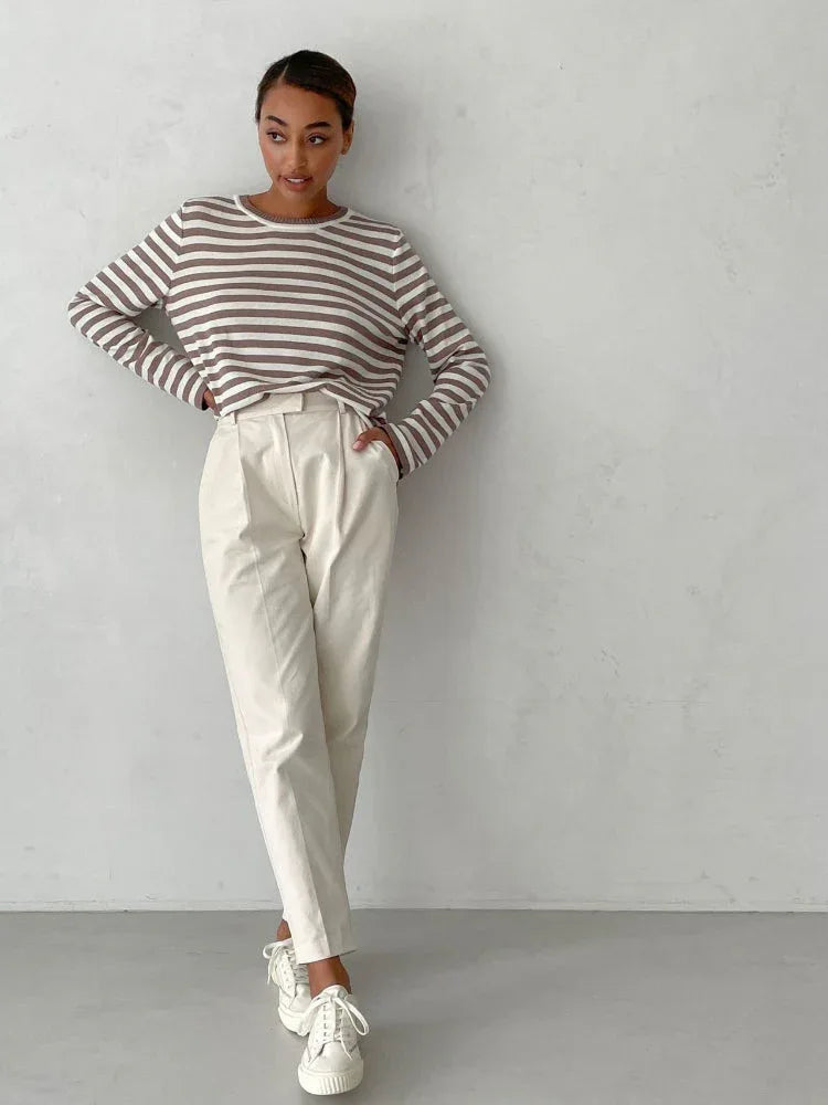 Full body shot of a woman wearing a brown and white striped long-sleeved crew neck sweater with cream-colored pants and white sneakers, hands on her hips.