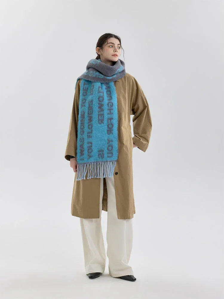 A woman models a blue and gray knit scarf with text print and fringe detail, worn over a tan trench coat and cream pants.