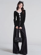 Full body shot of a woman wearing a black long-sleeved top with white lace trim and decorative buttons, dark wide-leg jeans, and carrying a black quilted handbag.