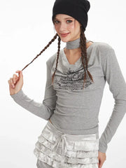 Close-up of a model's upper body and face wearing a grey long-sleeved top with a graphic print and choker detail, black beanie, playing with her braided hair.
