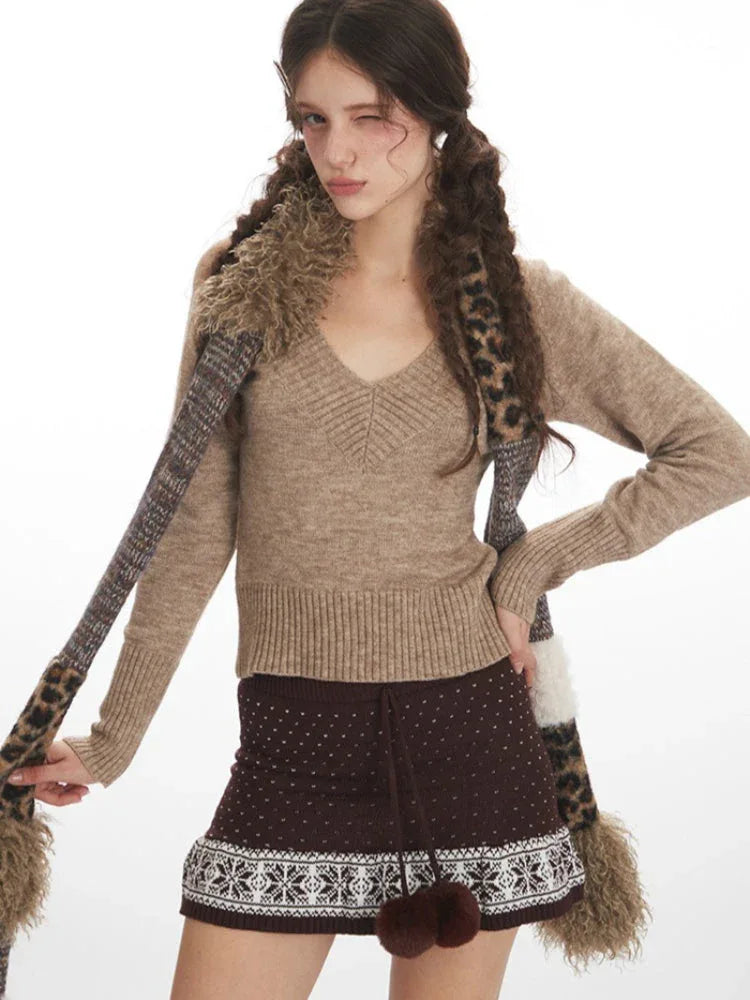 Close-up front view of a woman wearing a beige V-neck sweater, brown patterned mini skirt, and a patterned scarf, posing with one hand on her hip.