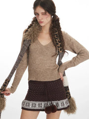 Close-up front view of a woman wearing a beige V-neck sweater, brown patterned mini skirt, and a patterned scarf, posing with one hand on her hip.