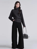 Woman wearing a dark grey turtleneck sweater with batwing sleeves and a cinched button detail at the waist, paired with black wide-leg pants and a silver handbag.
