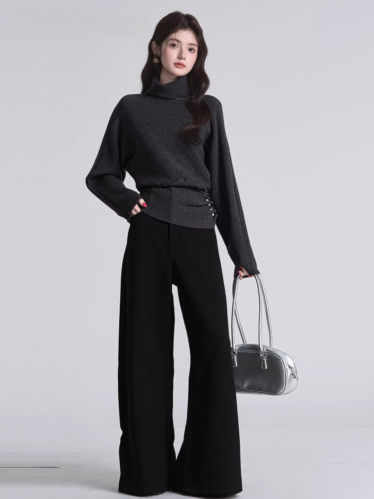 Woman wearing a dark grey turtleneck sweater with batwing sleeves and a cinched button detail at the waist, paired with black wide-leg pants and a silver handbag.
