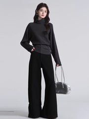 Woman wearing a dark grey turtleneck sweater with batwing sleeves and a cinched button detail at the waist, paired with black wide-leg pants and a silver handbag.