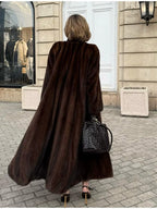 Back view of a woman wearing a long, brown mink fur coat, black leggings, black woven handbag, and brown pointed-toe heels, walking on a cobblestone street.
