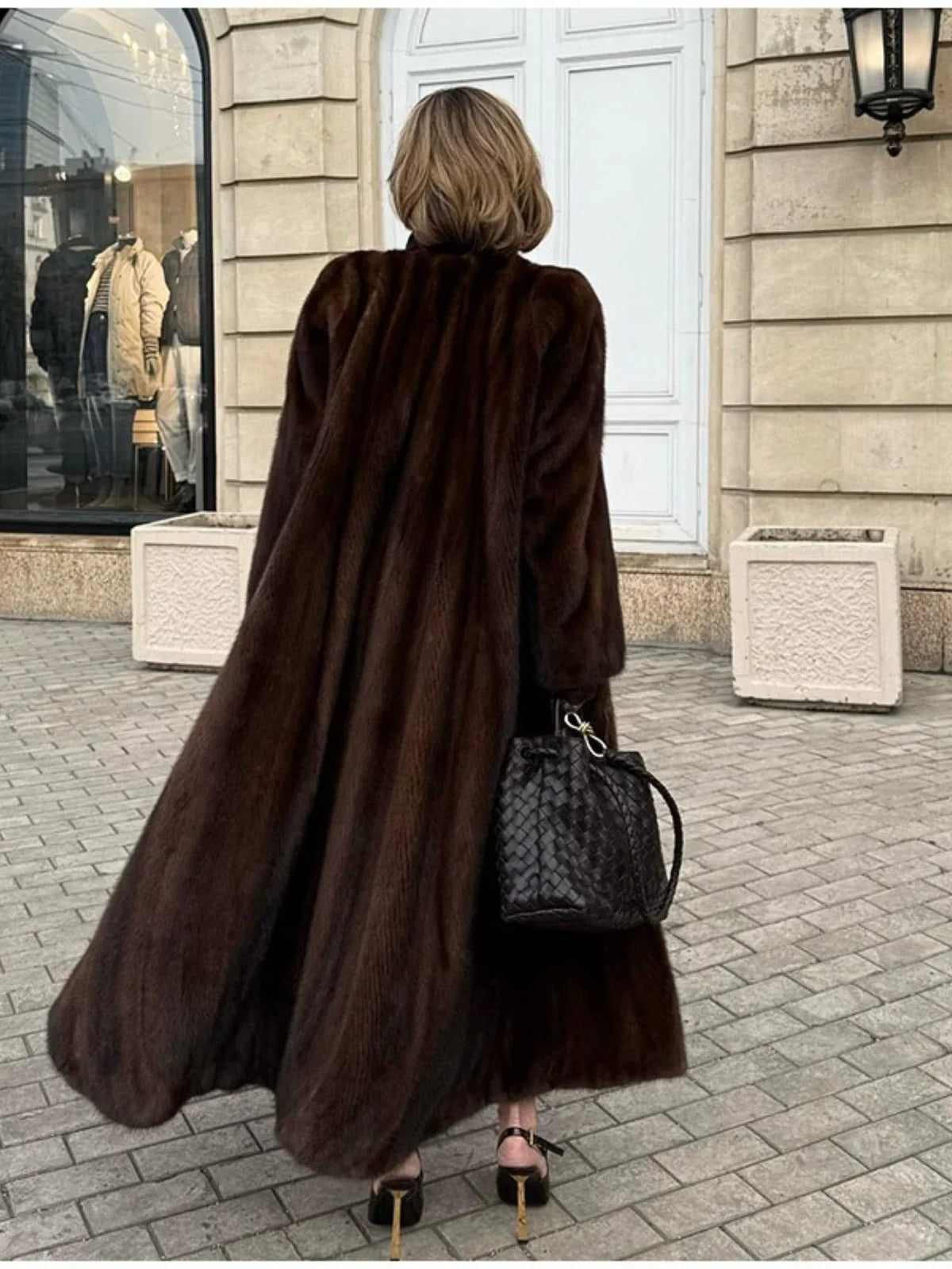 Back view of a woman wearing a long, brown mink fur coat, black leggings, black woven handbag, and brown pointed-toe heels, walking on a cobblestone street.