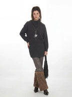 Woman wearing a charcoal grey oversized turtleneck knit tunic over patterned leggings and brown furry leg warmers, holding a black fringed handbag and wearing a star pendant necklace.