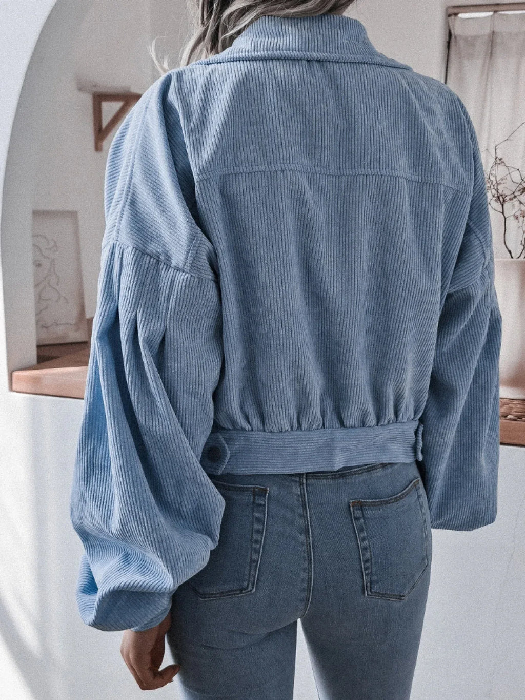Back view of a woman wearing a light blue corduroy cropped jacket with balloon sleeves and blue jeans.