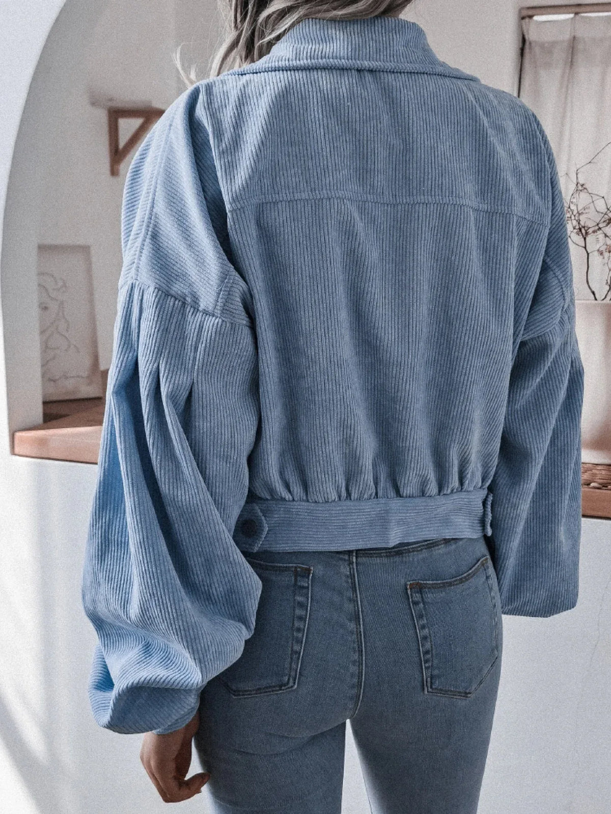 Back view of a woman wearing a light blue corduroy cropped jacket with balloon sleeves and blue jeans.