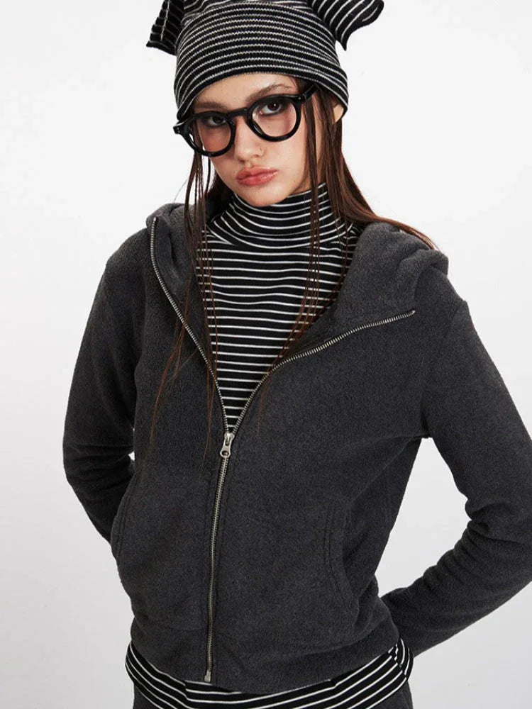 Close-up front view of a woman wearing a charcoal grey hooded zip-up jacket over a black and white striped turtleneck, looking to the side.