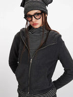 Close-up front view of a woman wearing a charcoal grey hooded zip-up jacket over a black and white striped turtleneck, looking to the side.