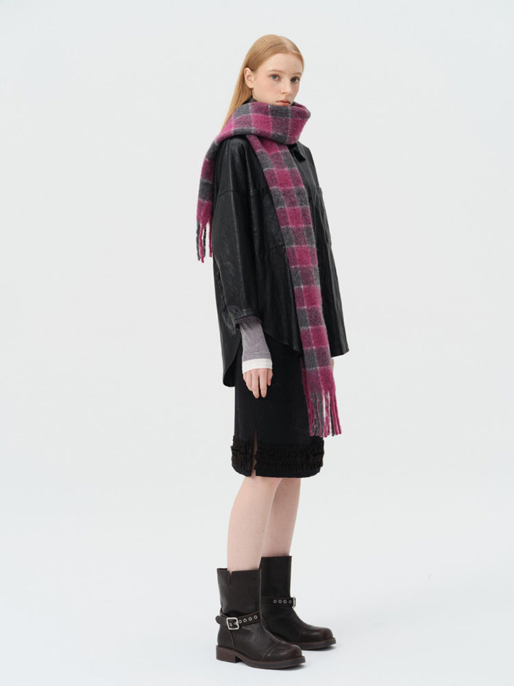 Three-quarter view of a woman wearing a black leather-look jacket, a black skirt with ruffled hem, a pink and grey plaid scarf, and black boots.