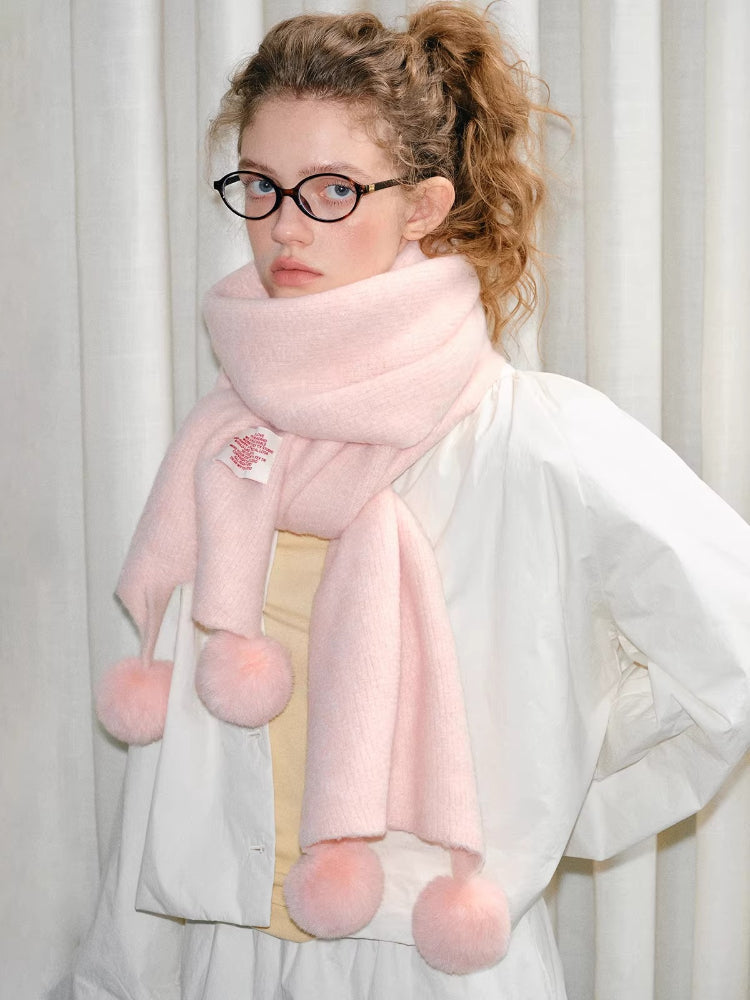 Woman wearing a pink fuzzy knit scarf with pom-pom embellishments, looking at the camera.