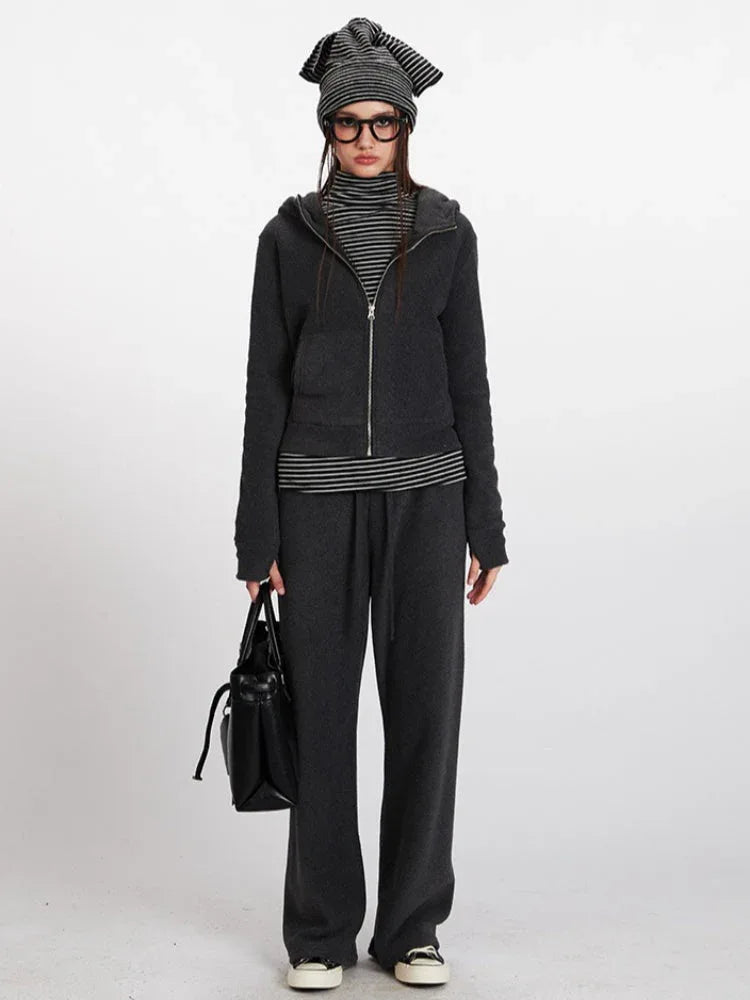 Full-length shot of a woman wearing a charcoal grey hooded zip-up jacket, matching wide-leg pants, a black and white striped turtleneck, and a striped beanie.