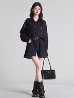 Full body shot of a woman in a dark wash denim shirt dress with a brown belt, holding a black handbag and wearing black boots.