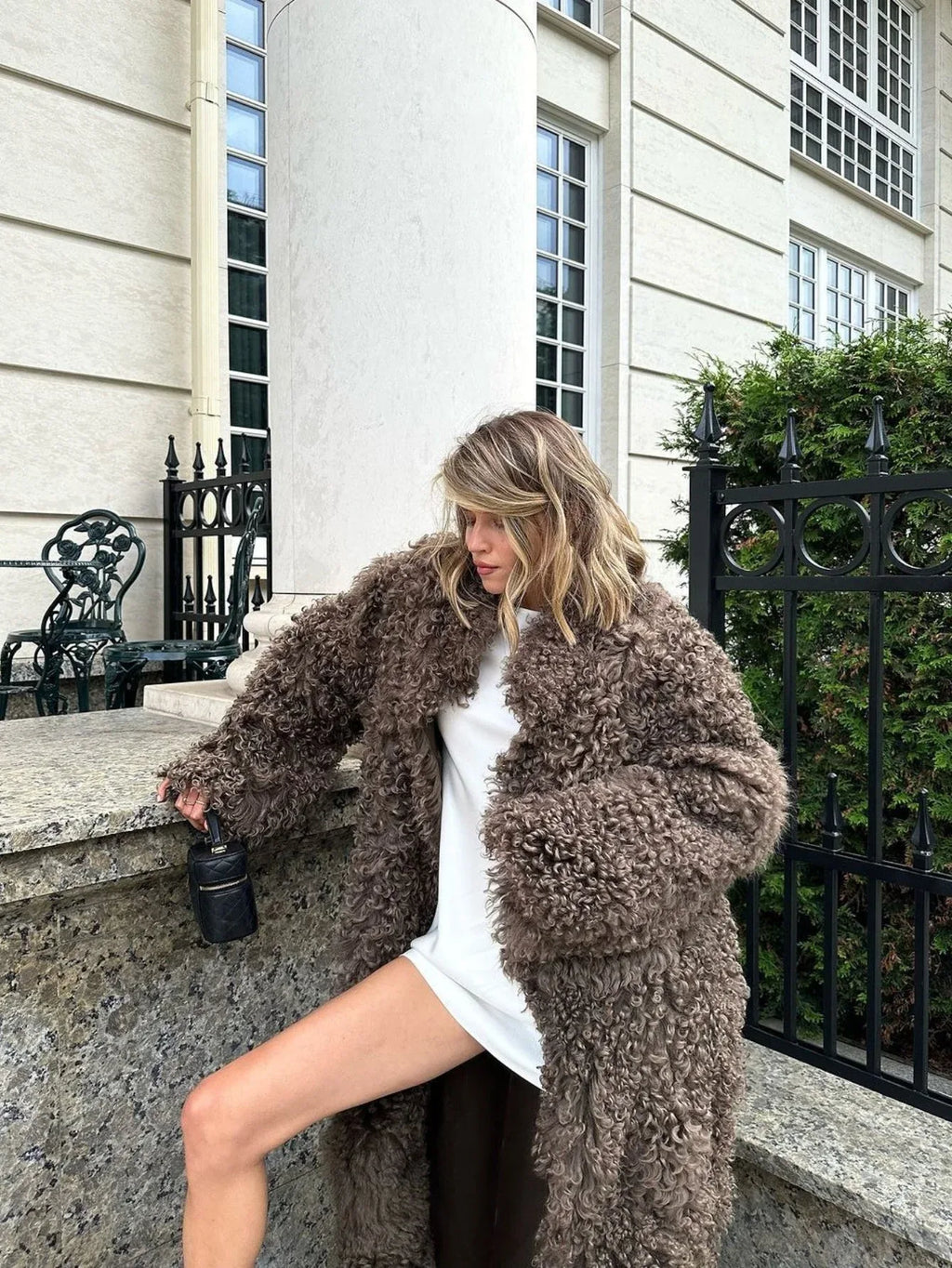 Woman wearing a long, fluffy brown faux fur coat over a white dress, holding a black handbag.