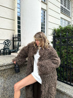 Woman wearing a long, fluffy brown faux fur coat over a white dress, holding a black handbag.