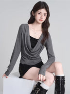 Woman wearing a grey draped long-sleeve top over a black camisole, paired with black shorts and black boots, sitting on a white cube.