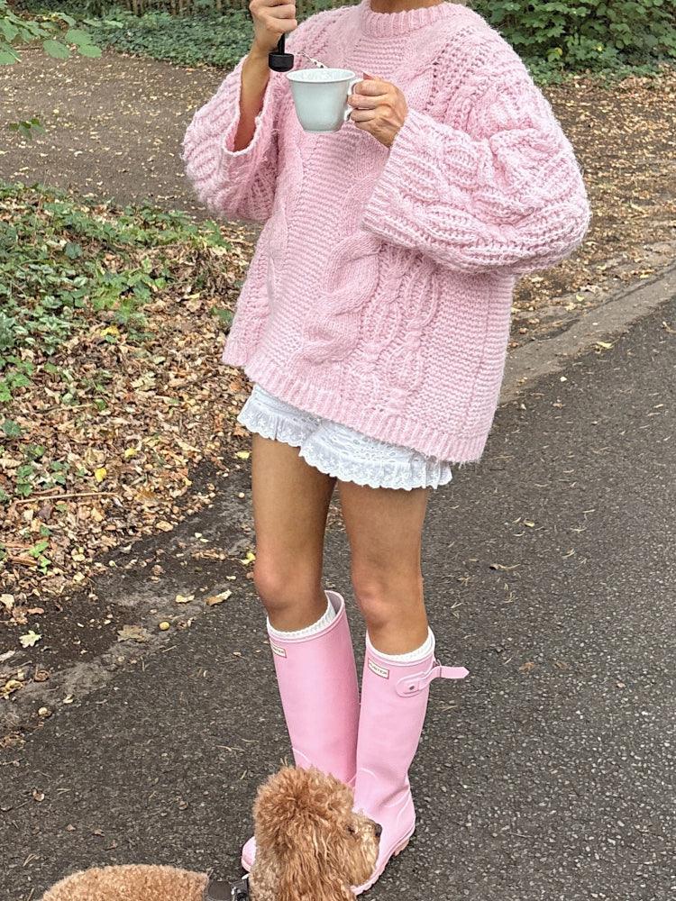 Woman in a pink oversized cable knit sweater, white shorts, and pink rain boots, holding a mug and an umbrella, with a poodle.
