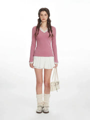 Woman wearing a pink long-sleeved top with white lace trim and white pleated shorts, holding a cream handbag and wearing cream leg warmers and silver shoes.