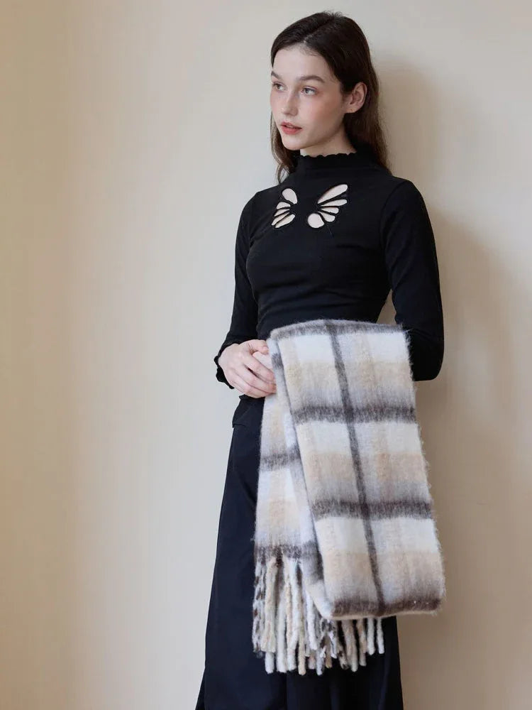 Close-up of a woman wearing a black mock-neck top with cut-out details, holding a folded plaid scarf with fringed edges.