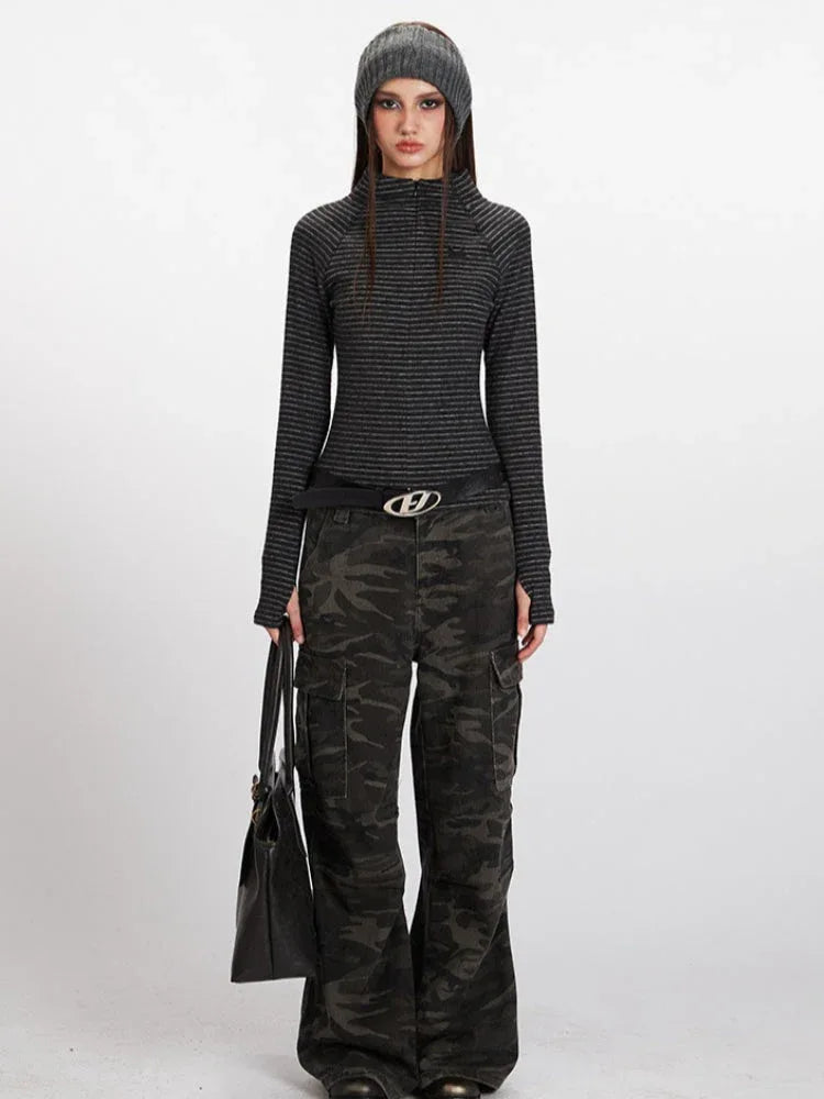 Model wearing a gray striped long-sleeve top with a mock neck and thumbholes, paired with camouflage cargo pants and a black belt, holding a black tote bag.