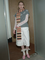 A woman stands wearing a gray sweater with lace sleeves, white shorts, pink shoes, and a striped scarf.