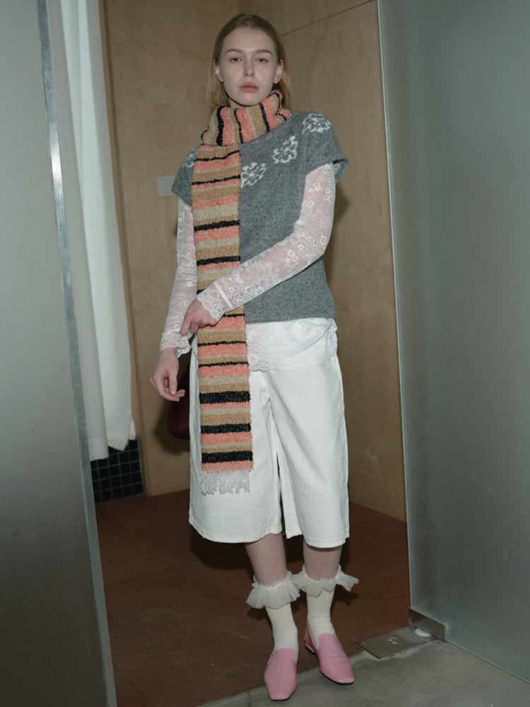 A woman stands wearing a gray sweater with lace sleeves, white shorts, pink shoes, and a striped scarf.