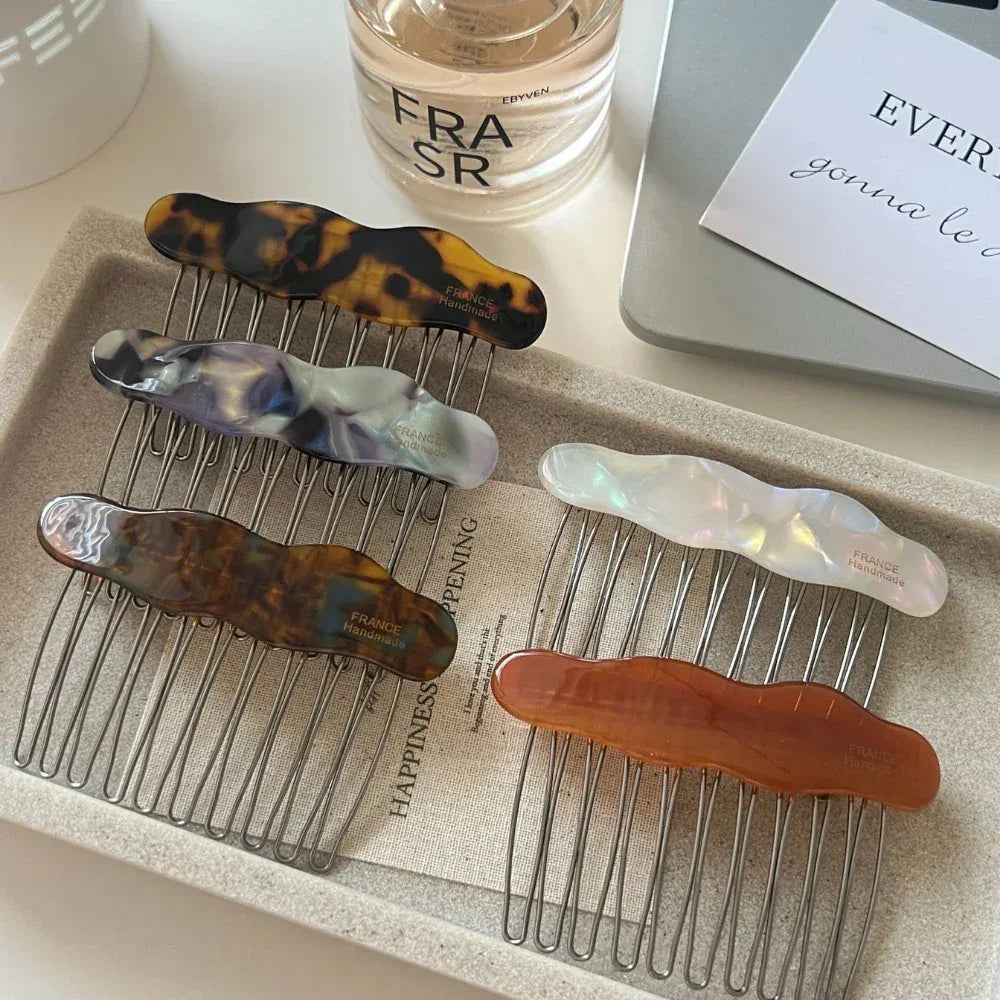 A collection of cloud-shaped hair combs in various colors and patterns, including tortoiseshell, marbled purple and grey, white iridescent, brown and blue marbled, and amber, arranged on a tray.