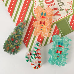 An overhead view of four Christmas-themed hair claw clips: a green Christmas tree, a striped candy cane, an orange gingerbread man, and a teal gingerbread man, laid out on a festive background.