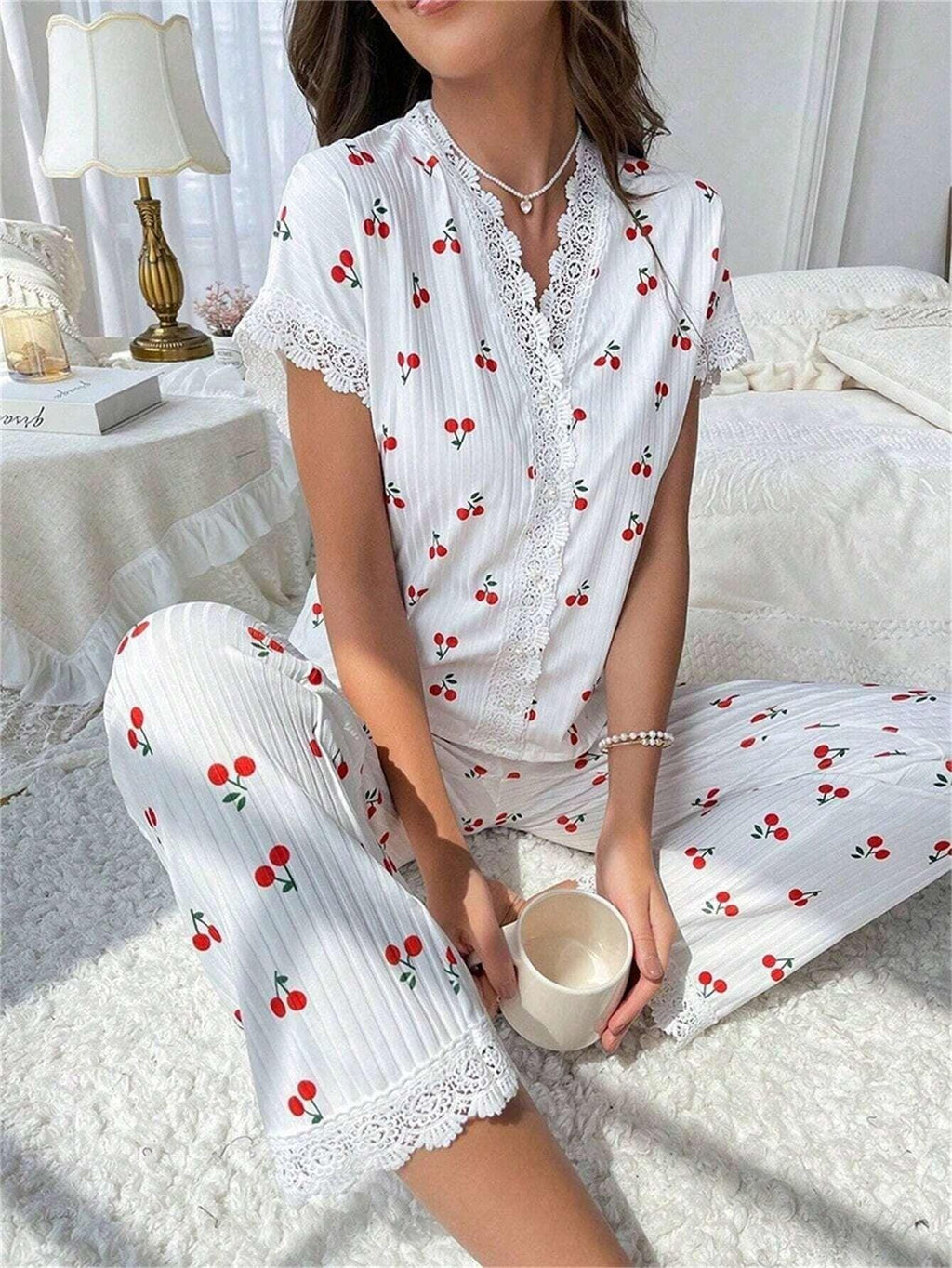 Woman wearing white short-sleeve pajama top with cherry print and white ribbed pants with cherry print, sitting on the floor in a bedroom, holding a mug.