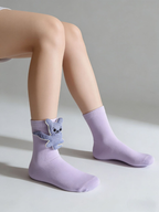 A person wearing light purple ankle socks with blue knitted bear embellishments on each sock, shown from the knees down.