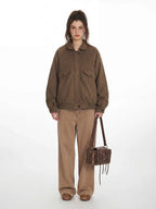 Woman wearing a brown bomber jacket and tan pants, holding a brown bag.