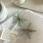 Two starfish hair clips, one light blue and one light grey, are placed on white crumpled paper.