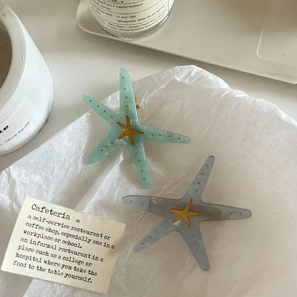 Two starfish hair clips, one light blue and one light grey, are placed on white crumpled paper.