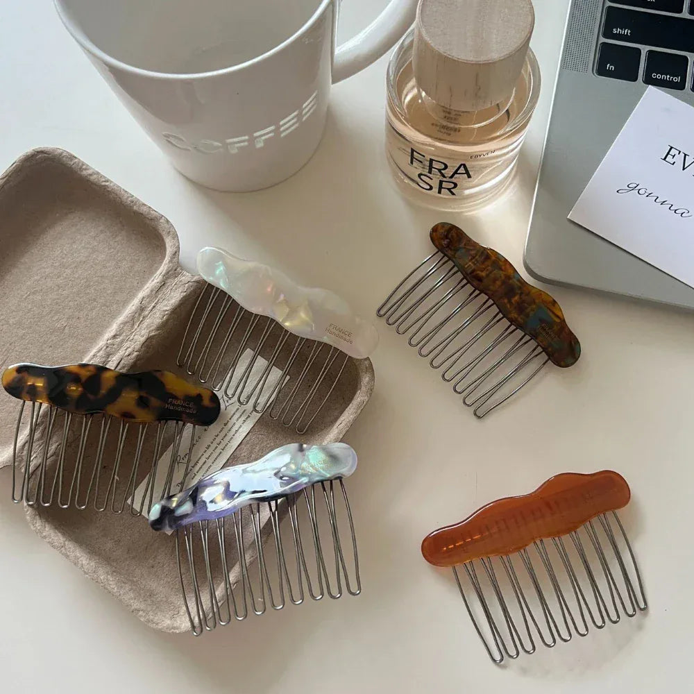 Several cloud-shaped hair combs in different colors are laid out on a white table, with a coffee mug, a perfume bottle, and a laptop visible.