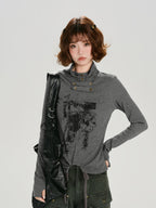 Woman wearing a grey long-sleeve graphic print turtleneck top with thumbhole cuffs and a drawstring hem, with a black bag slung over her shoulder.